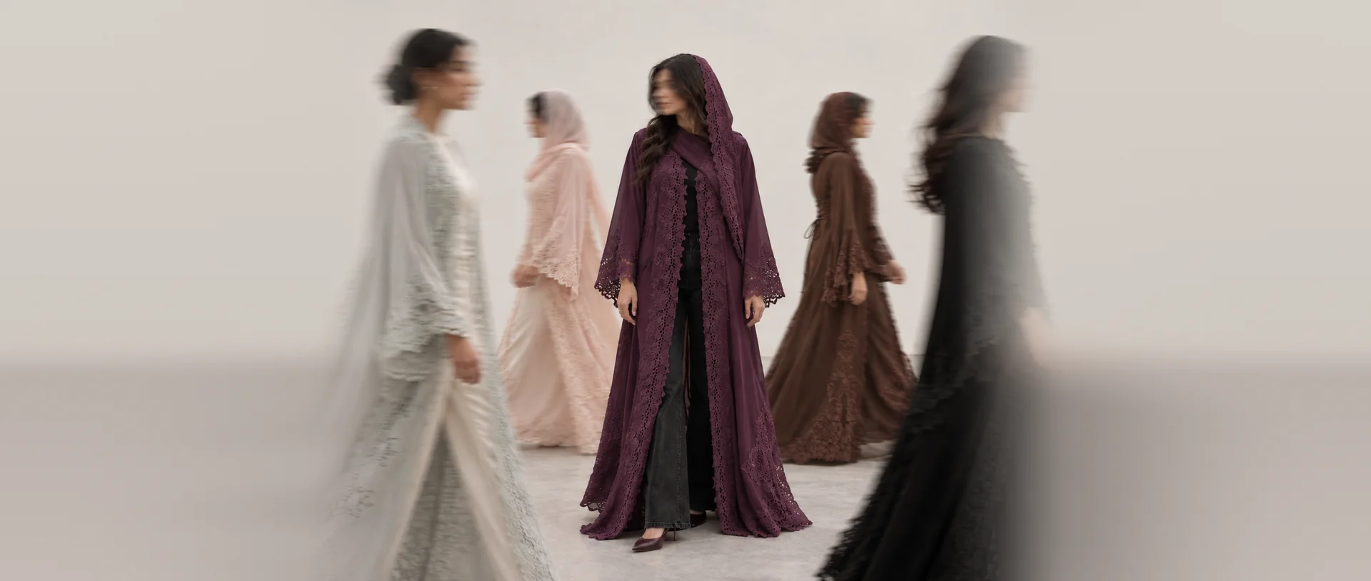 In a pale studio, a woman in Layali, deep aubergine lace with its hood up, stands still and in focus while four women in other colours walk past her, blurred with movement.