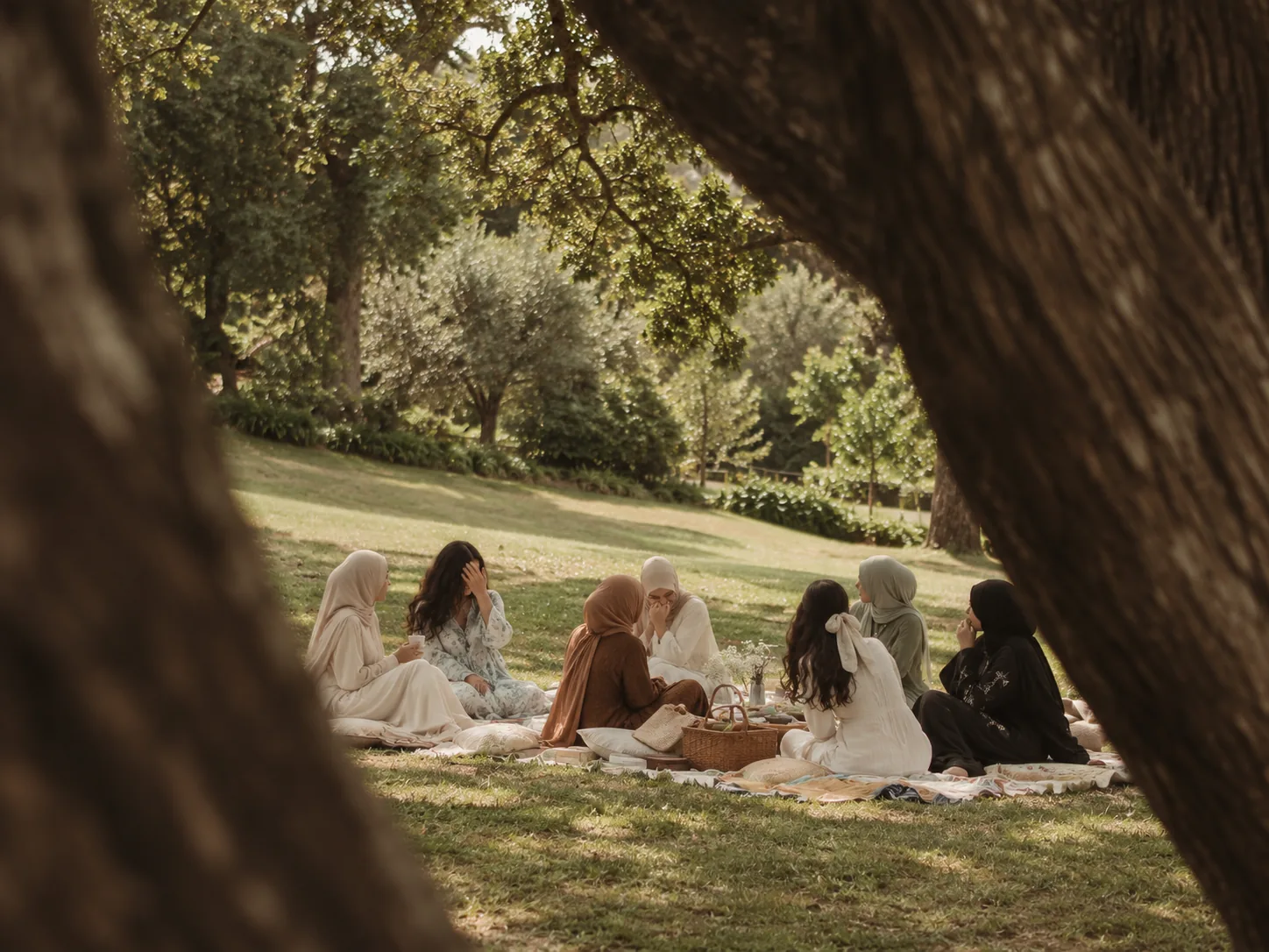 Seven women sitting together in a circle on blankets in a sunlit park, seen between two old trees, talking and laughing over a picnic basket and a jar of flowers.