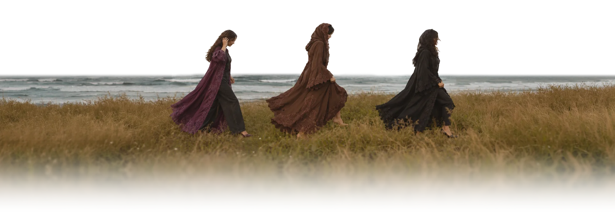 Three women walking together through tall grass by the sea, in Layali&rsquo;s deep aubergine, Tamr&rsquo;s espresso brown and Kohl&rsquo;s black, their abayas lifting as they go.